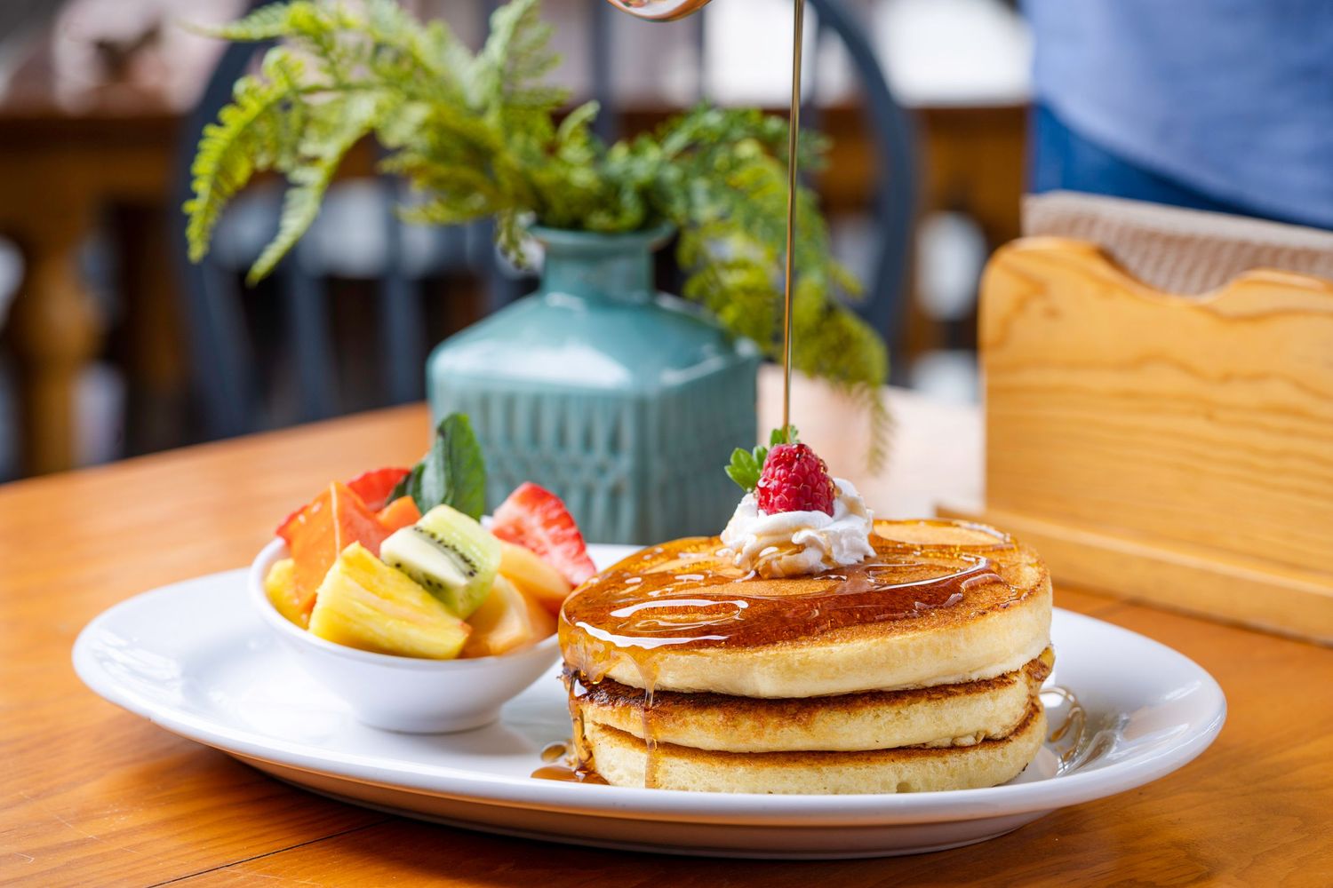 Stack of golden pancakes with syrup and butter on a white plate in a restaurant setting.