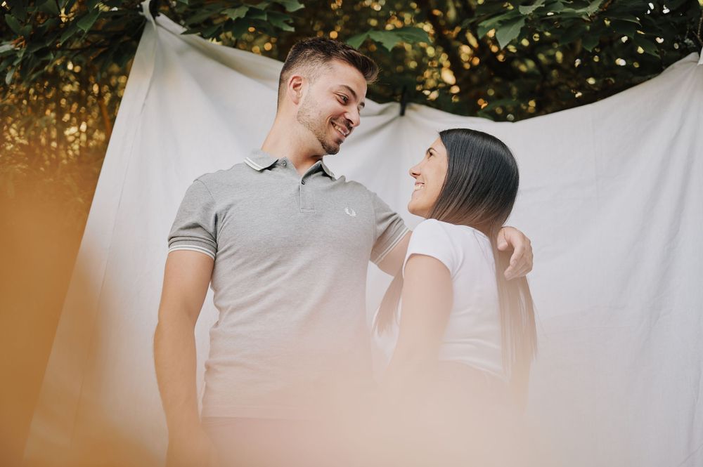 Smiling couple gazing at each other standing against white fabric backdrop in natural light.
