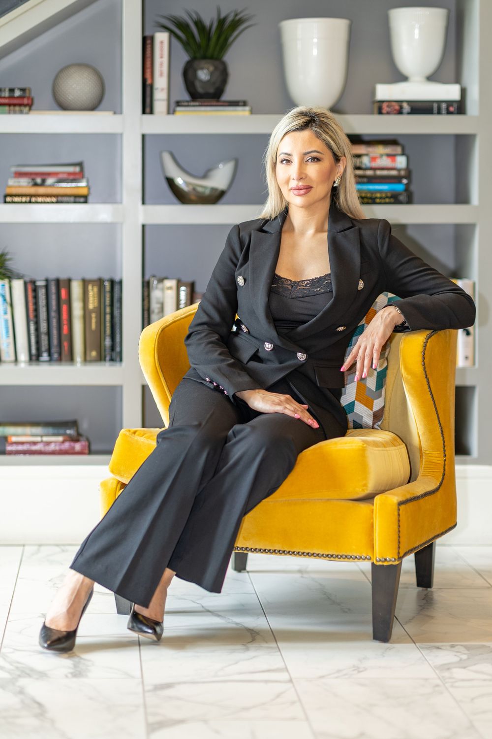 Professional in a black business suit relaxes in a yellow armchair against stylish built-in shelving with books and decor.