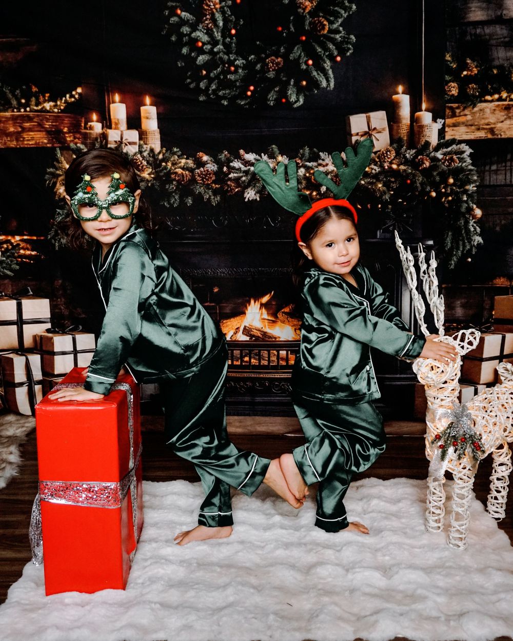 Two children in green satin Christmas pajamas pose by a decorated fireplace with gifts and a reindeer decoration.