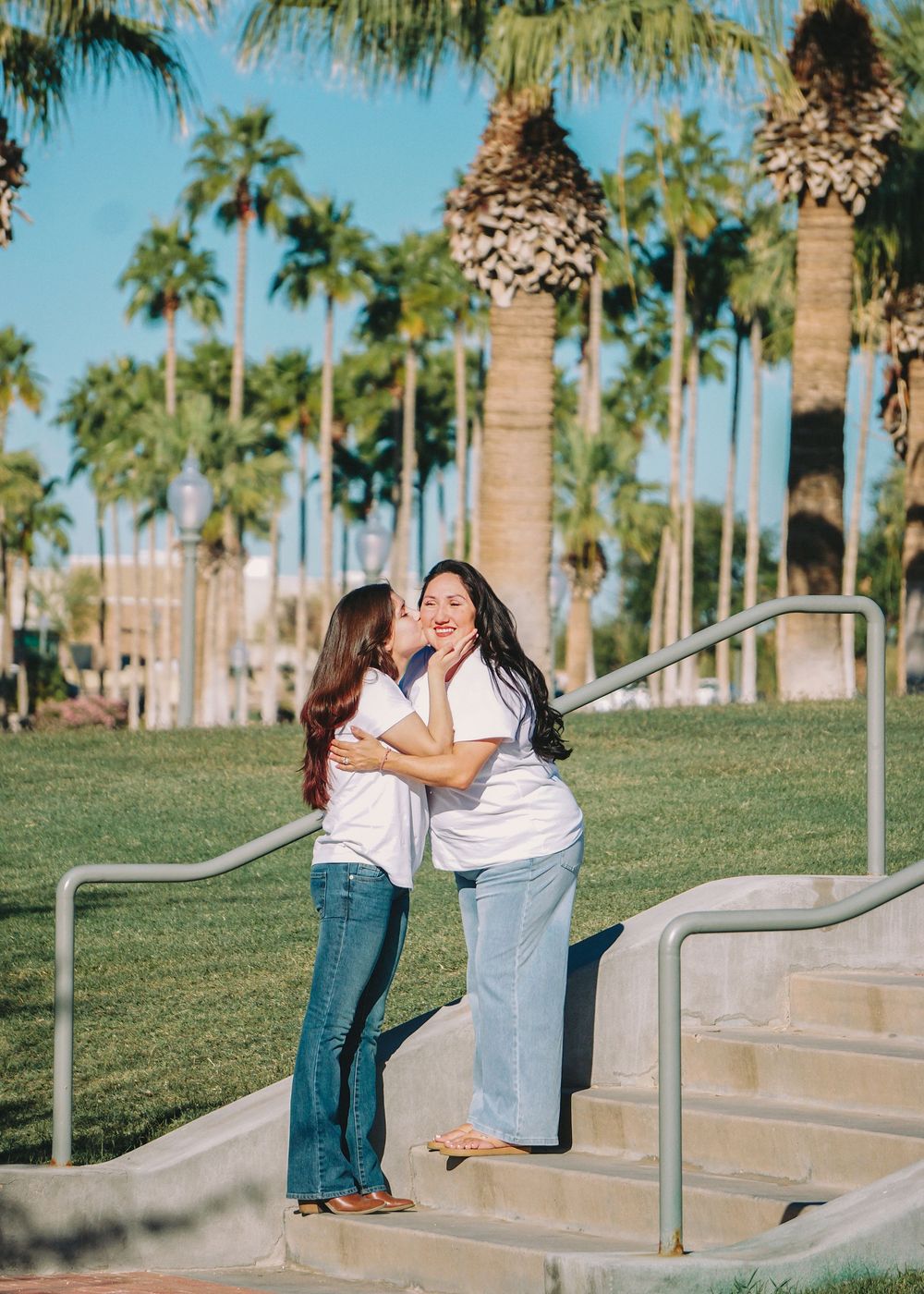Two women in white shirts embrace and share a kiss on outdoor steps surrounded by tall palm trees on a sunny day.