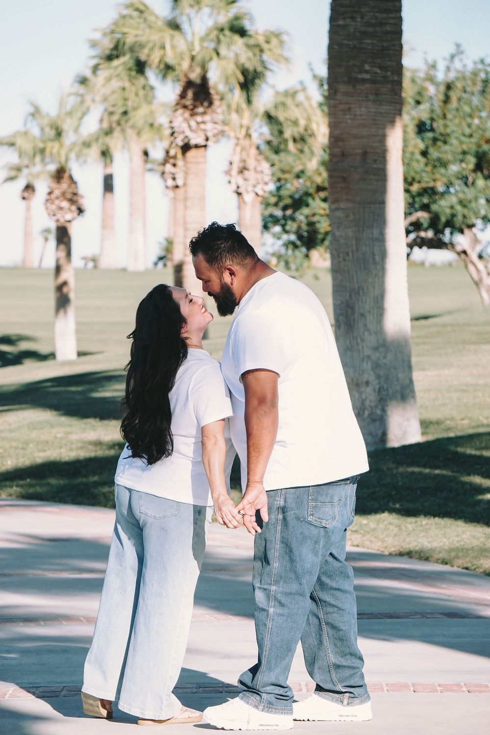 Couple sharing a romantic kiss in a sunny park with palm trees, both wearing white tops and jeans.