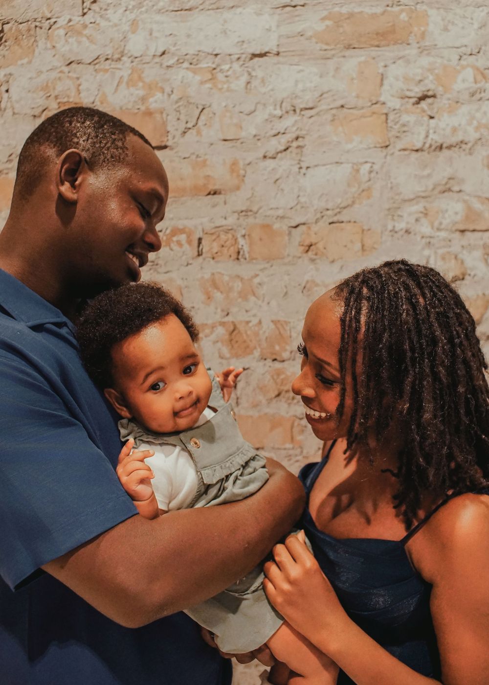 A happy Black family with a man and woman smiling at their baby against a rustic brick wall backdrop.