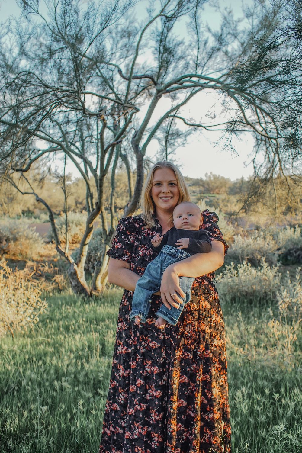 Woman in floral dress holds baby outdoors in sunny desert garden with trees.