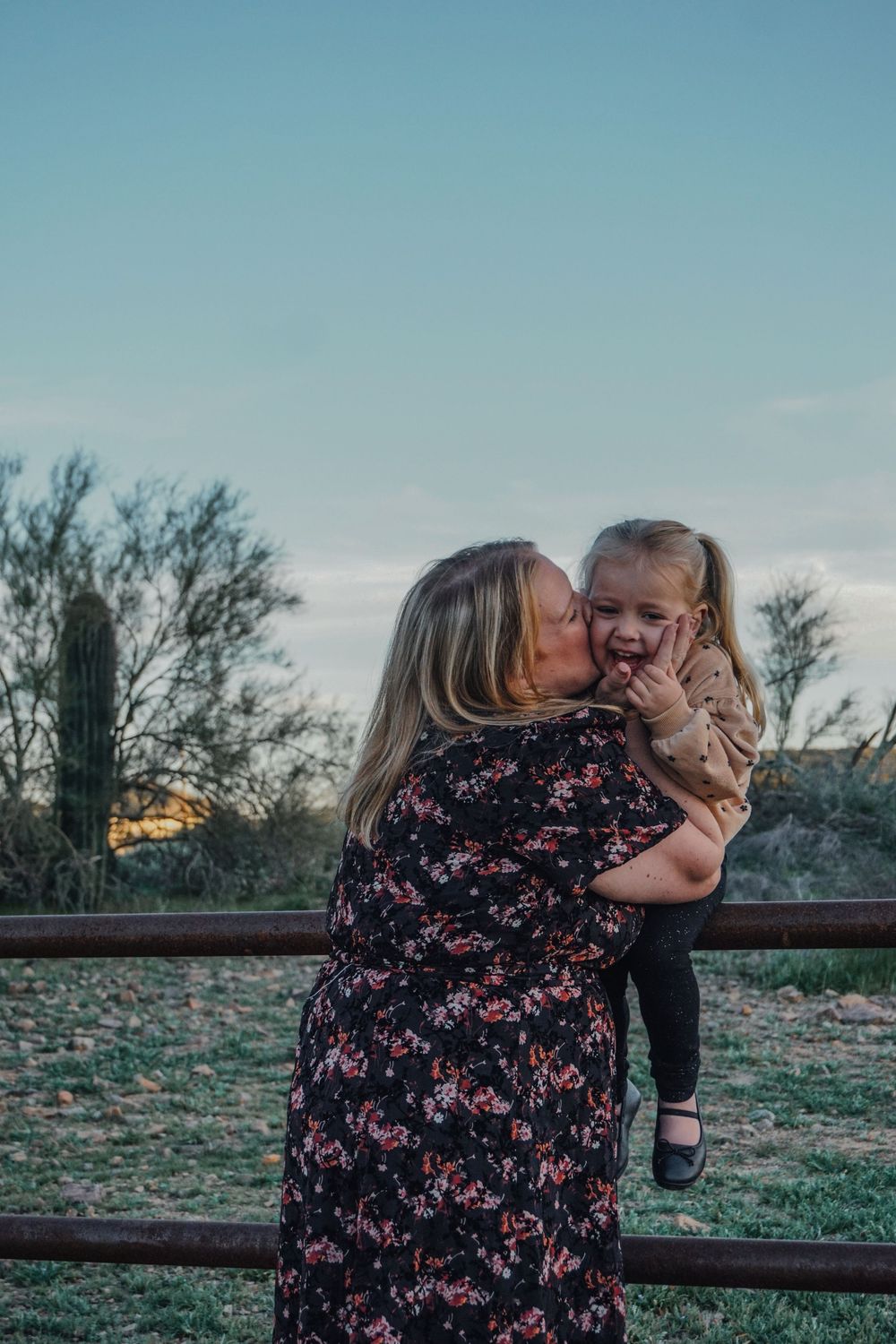 A woman in a floral dress kisses a laughing toddler girl outdoors at dusk near a wooden fence.