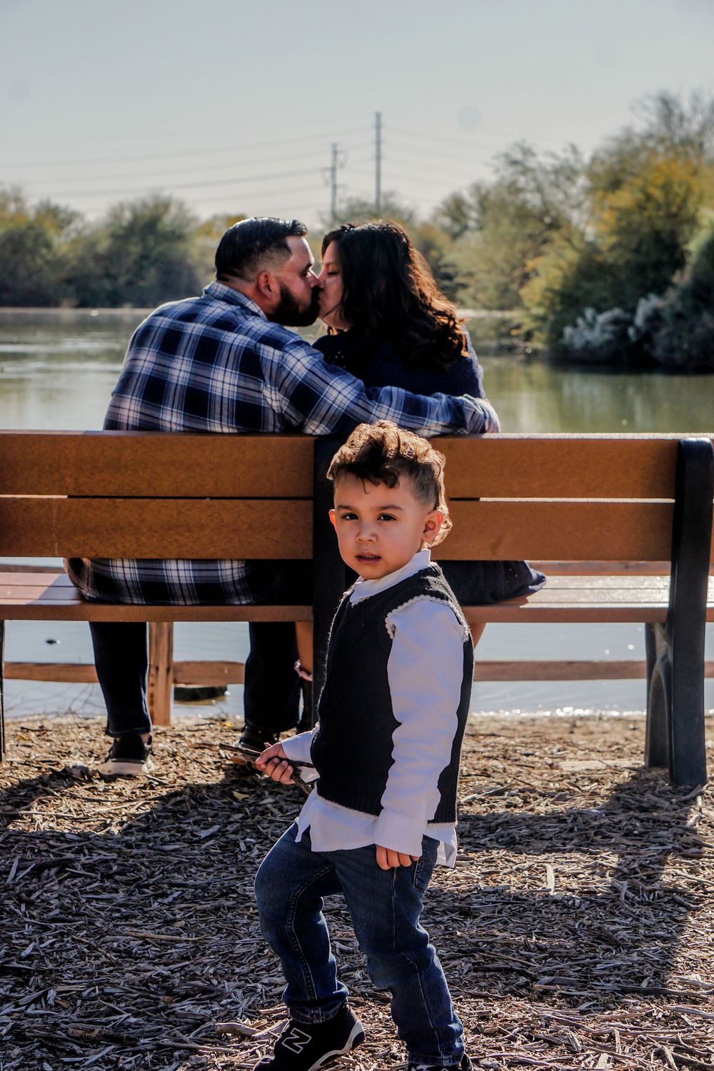 A young boy stands in front of a couple kissing on a bench by a lake outdoors.
