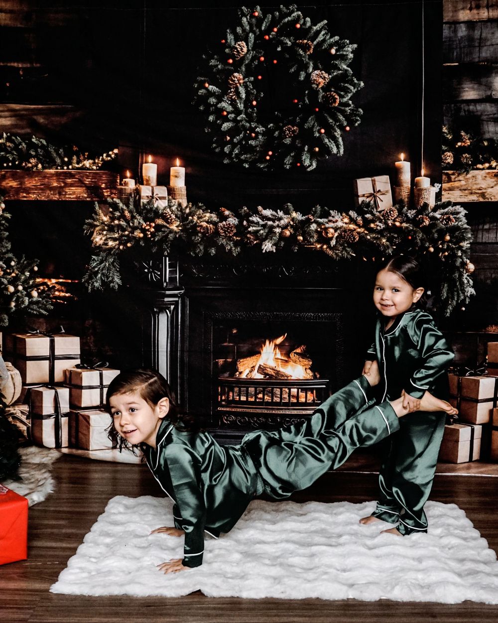 Two children in green satin pajamas pose playfully by a decorated fireplace with Christmas wreaths and gifts.