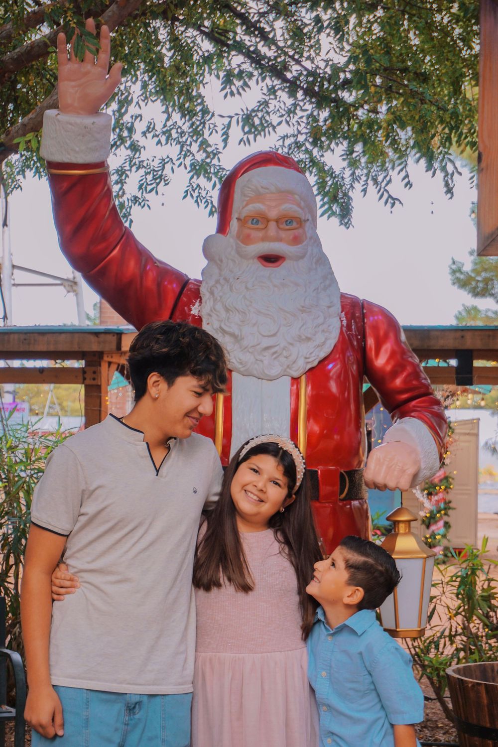 Three children pose happily in front of a large Santa Claus statue outdoors near festive holiday decorations.