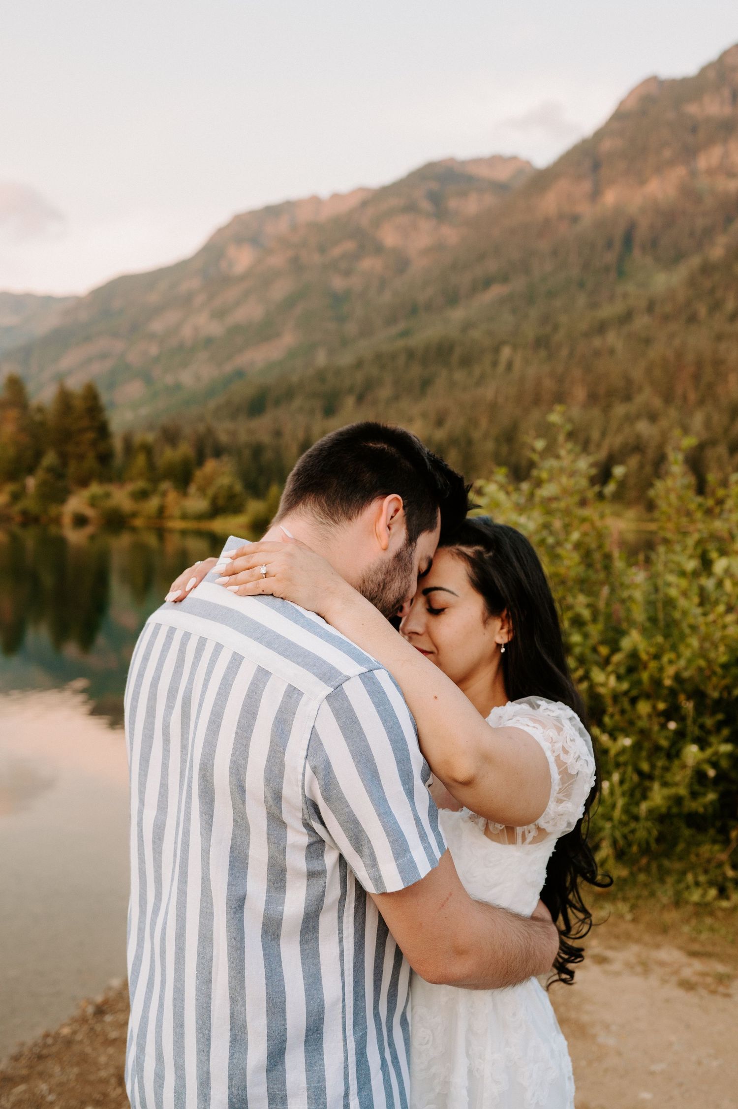Romantic embrace by a mountain lake during sunset with evergreen trees in background.