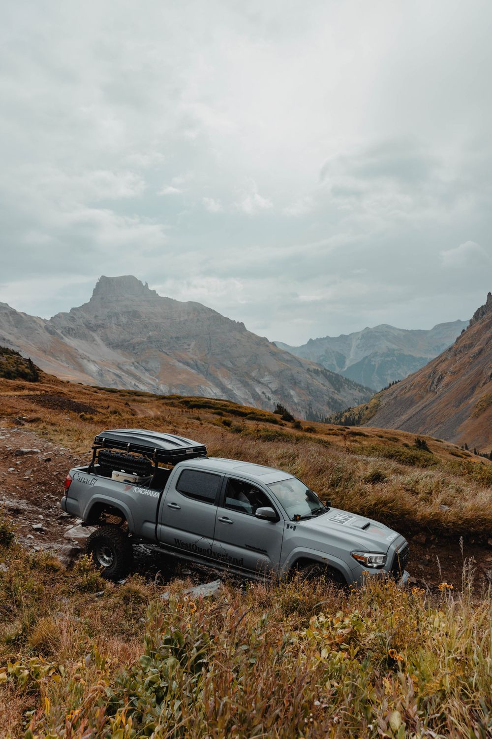 Gray pickup truck traversing mountain trail with dramatic alpine peaks in background.
