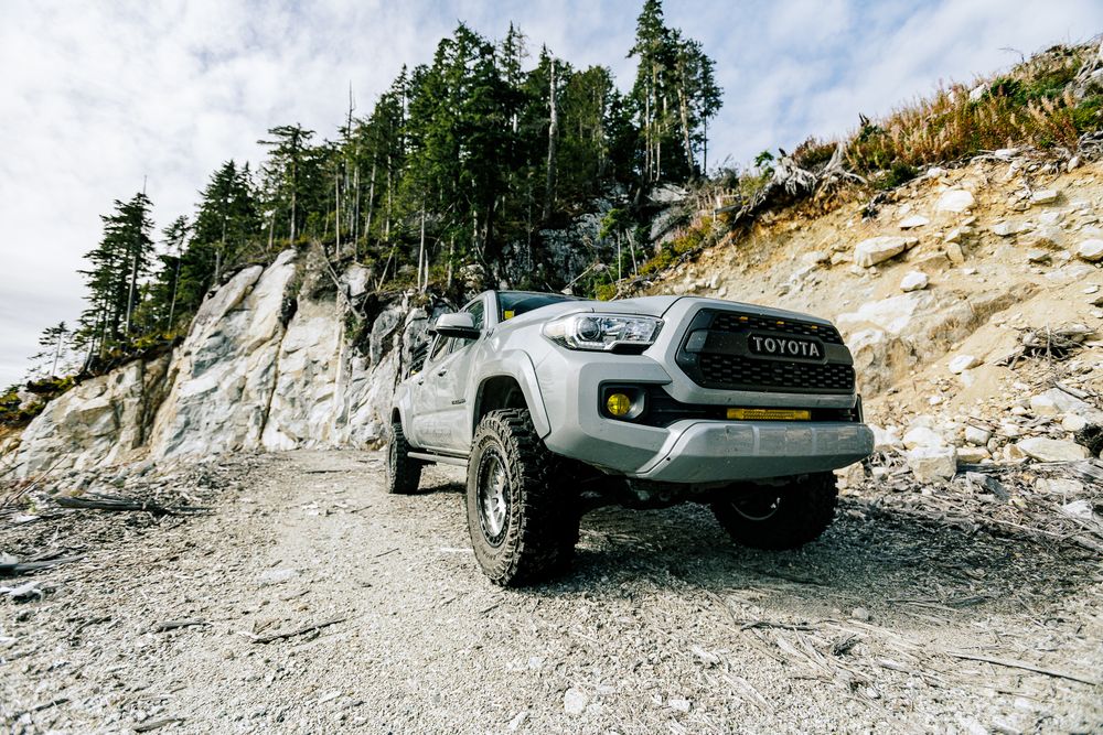 A silver SUV navigates along a narrow mountain road with steep rocky cliffs and evergreen trees in the background.