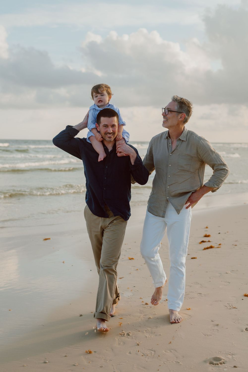 Three people enjoying a casual walk together on the beach at sunset with waves and cloudy sky in the background.