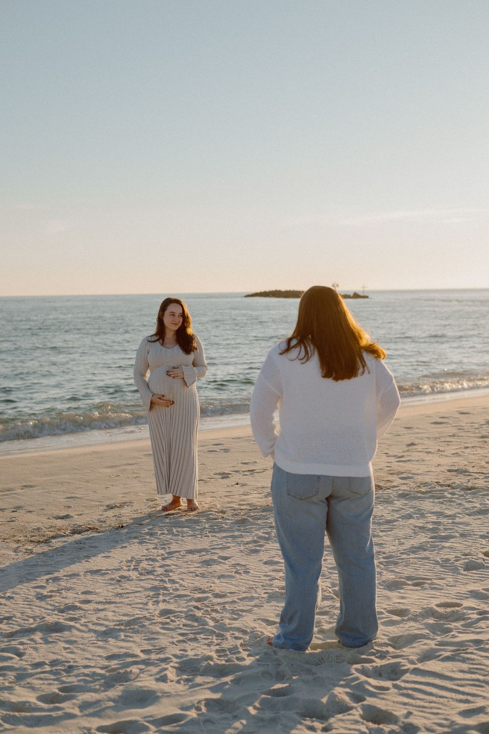 Two people in white tops and jeans walk along a serene beach at sunset with ocean waves in the background.