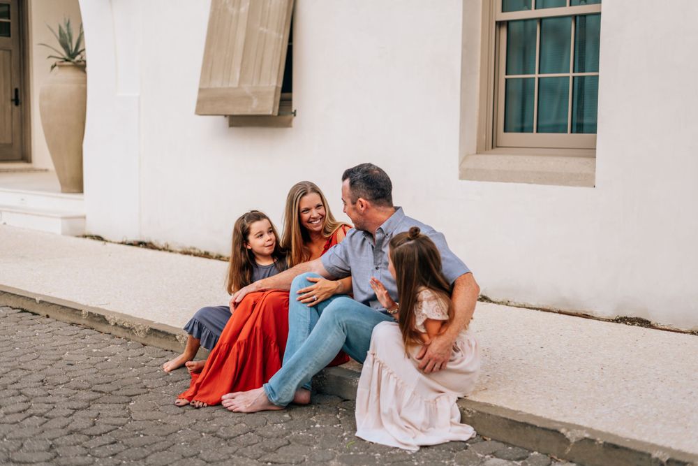Group sitting and laughing together on cobblestone street against white wall.
