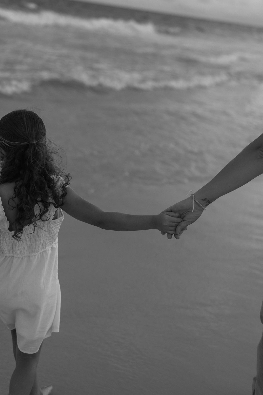 Black and white beach photo showing two people holding hands by the ocean waves, with one wearing a white summer dress.
