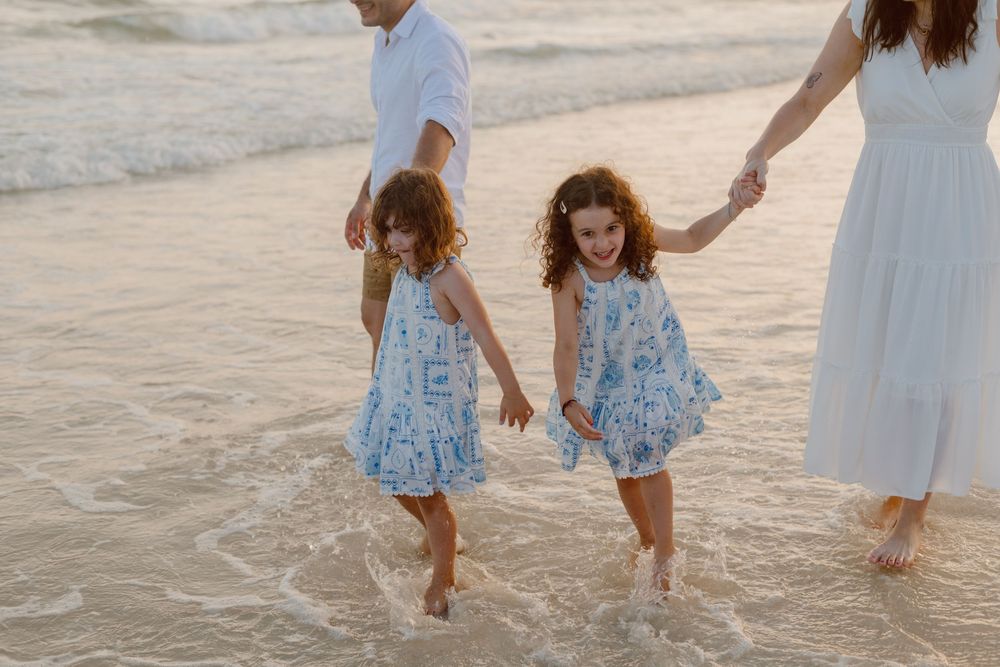 Several people wearing light colored clothes walk on a sandy beach at sunset, creating shadows on the wet sand.