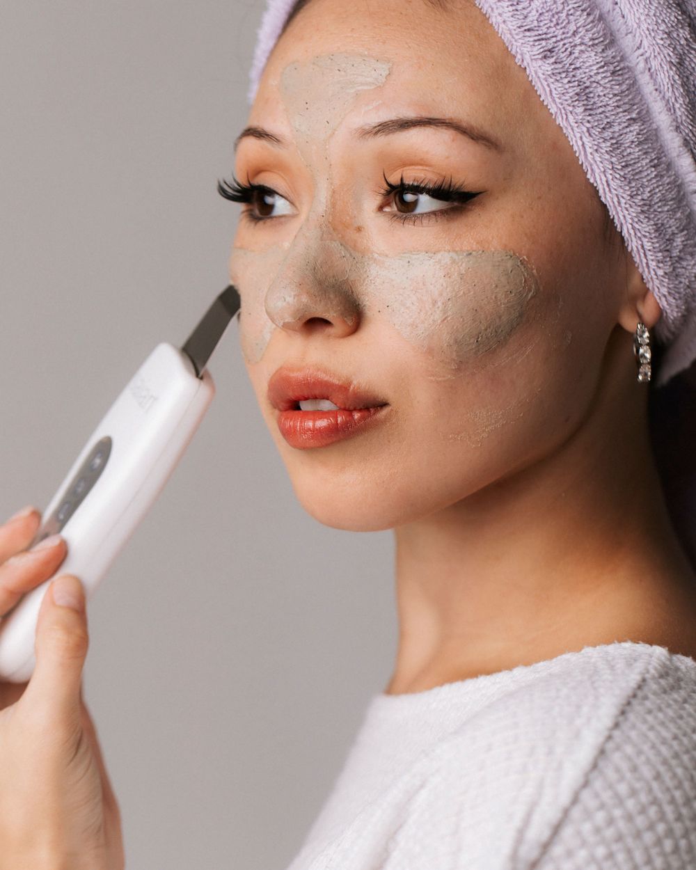 Woman applying clay face mask with ultrasonic skin scrubber device, hair wrapped in towel, skincare routine.