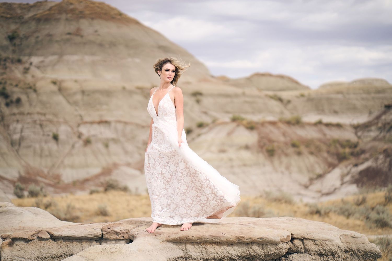 A figure in a flowing white lace dress stands gracefully on rocky terrain with eroded cliffs in the background on an adventure photo session