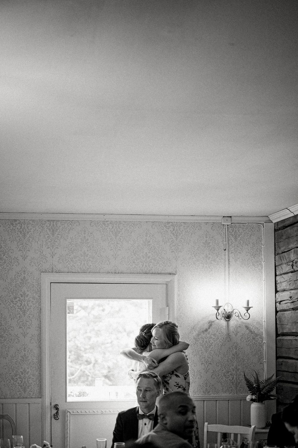 An intimate black and white indoor scene shows guests at what appears to be a wedding reception or celebration.