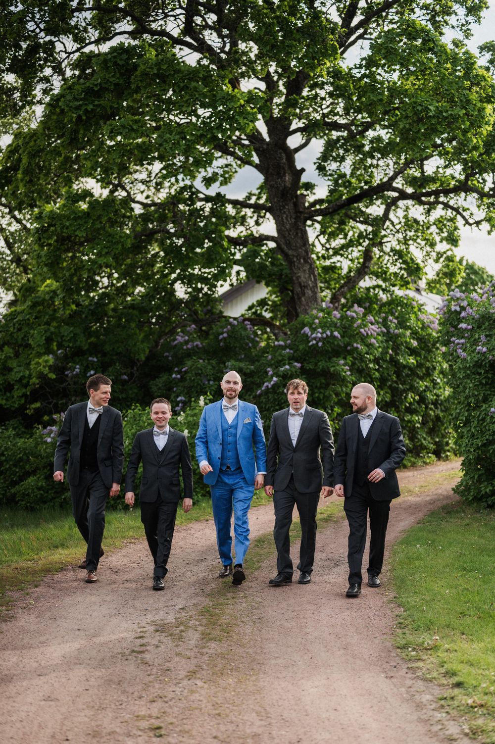 A group in formal attire walks together on a dirt path surrounded by green trees and nature.