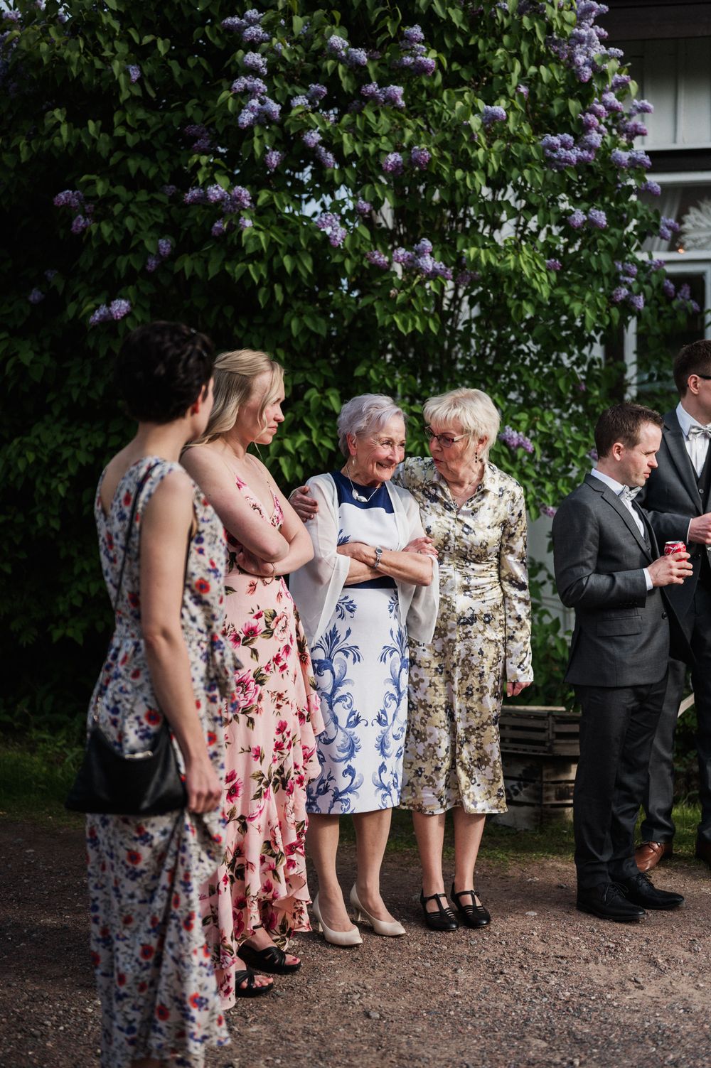 A group of wedding guests in floral dresses gather under blooming lilac bushes on a garden path.