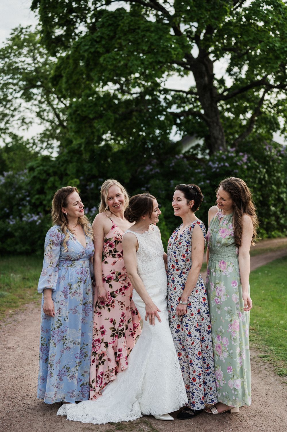 Wedding party wearing floral print maxi dresses in pastel colors stand together on a scenic garden path.