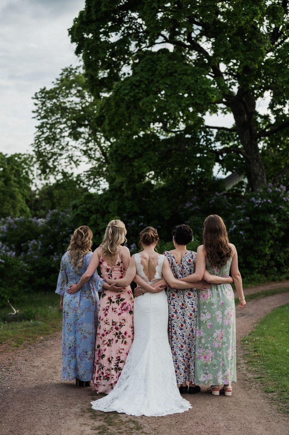 A bride and bridesmaids in floral maxi dresses stand together on a garden path with lush trees in the background.
