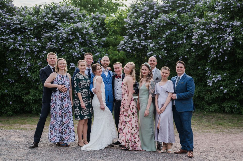 A wedding party poses together in front of flowering bushes wearing elegant dresses and suits.
