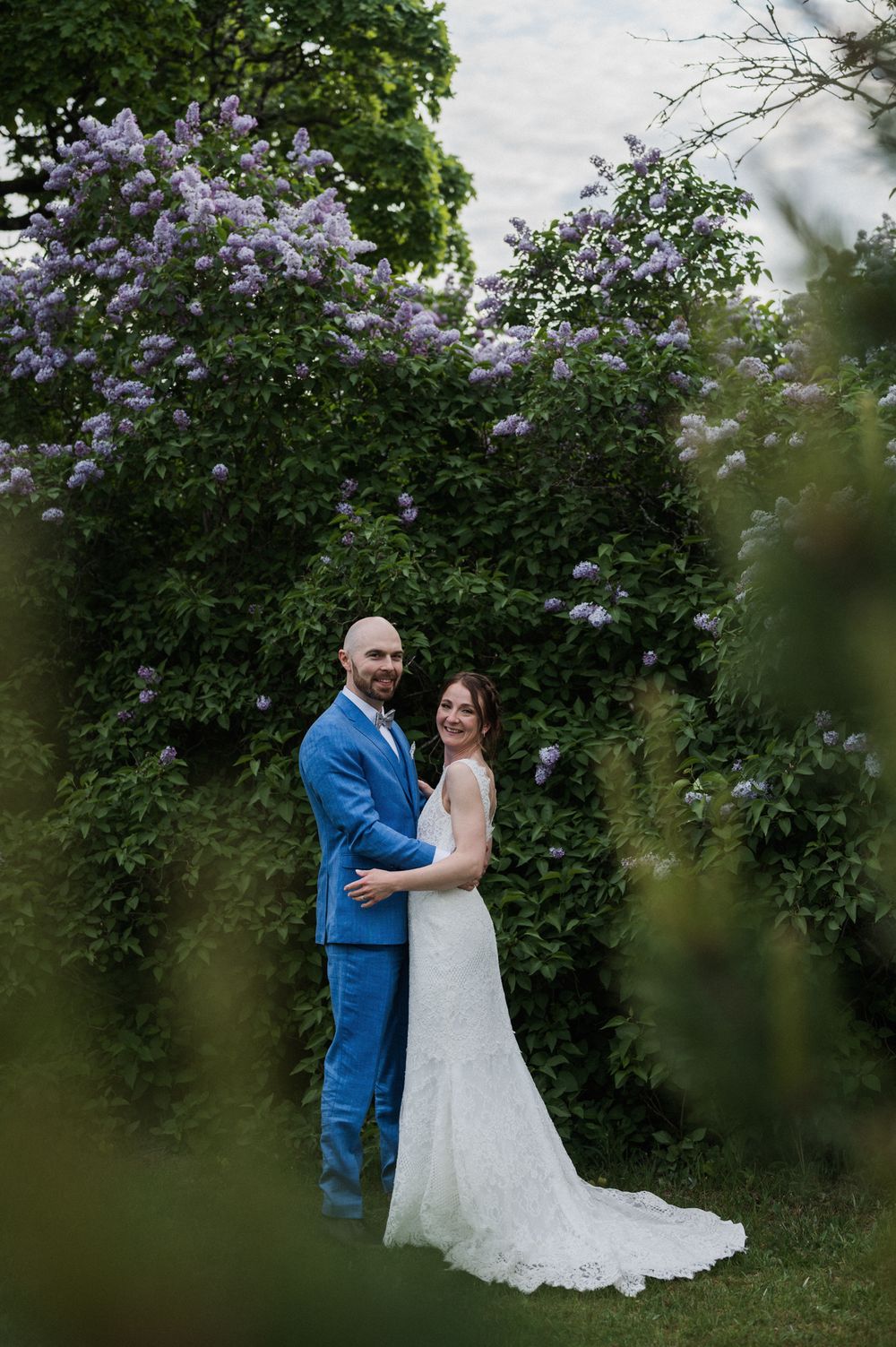 A couple poses for a romantic portrait against a backdrop of blooming purple lilac bushes.