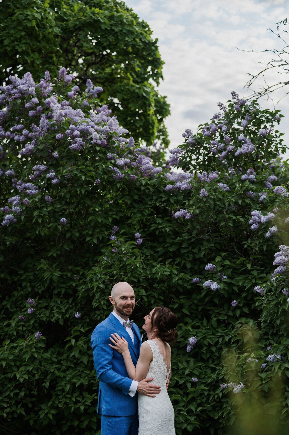 A couple shares an intimate moment in a garden with blooming purple lilacs and lush greenery.