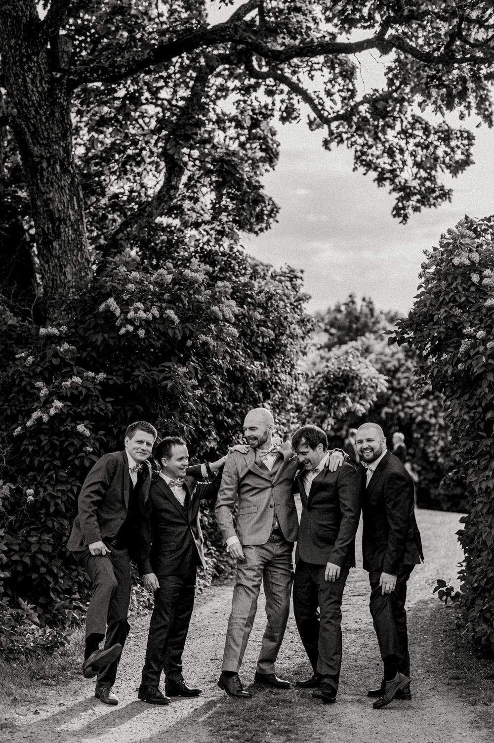 Group of five friends in formal attire share a candid moment on a tree-lined path in black and white.