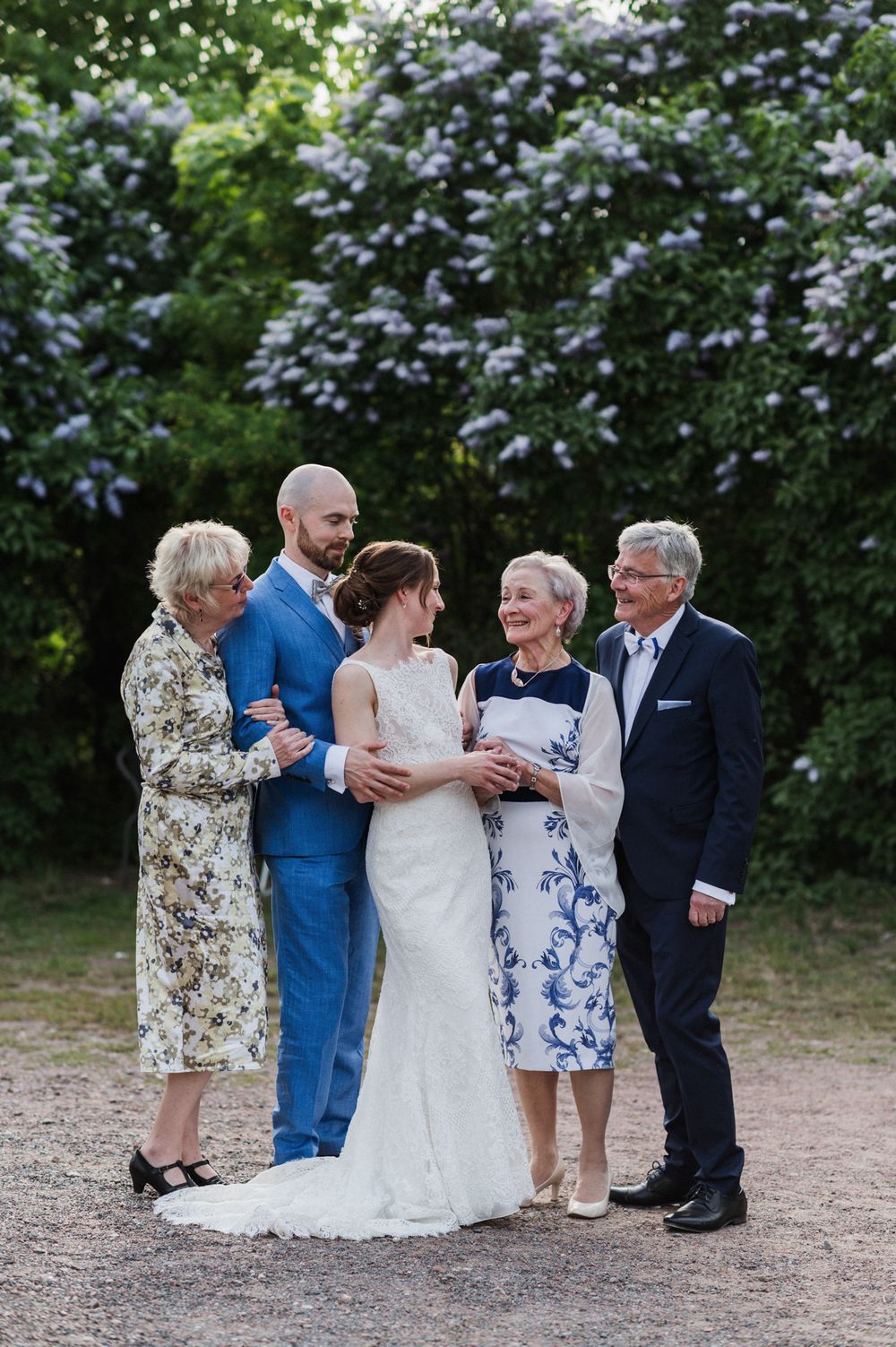A small wedding party poses together on a gravel path surrounded by flowering lilac bushes.