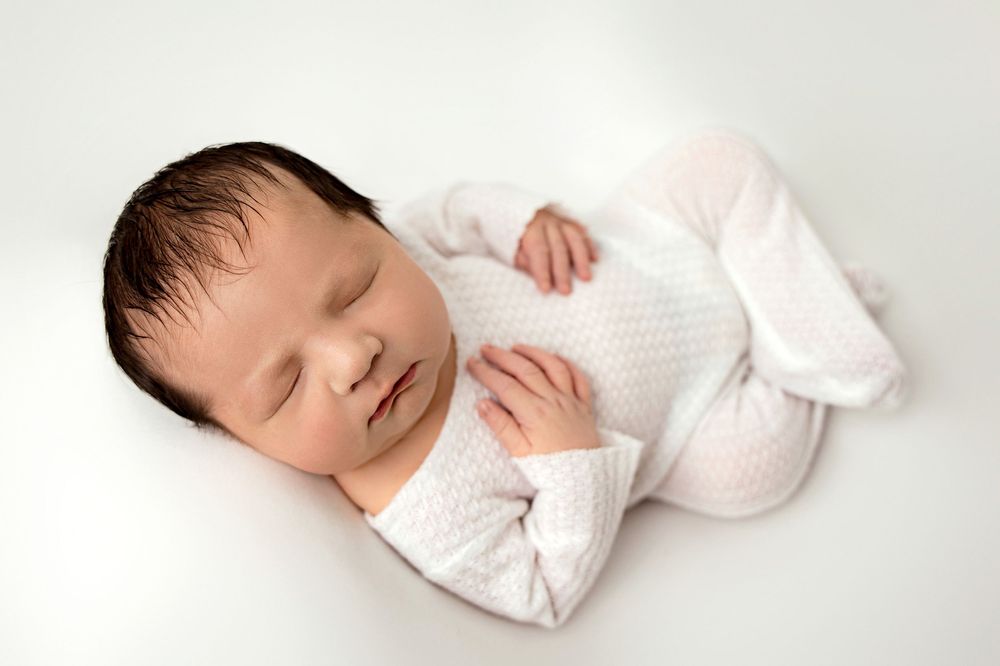 Sleeping newborn baby wearing pink headband and dress lying on white backdrop in studio setting.