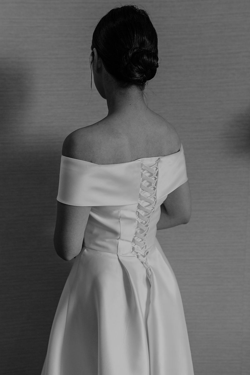 Back view of a white off-shoulder wedding dress with corset-style lacing detail and pleated skirt shown in black and white.