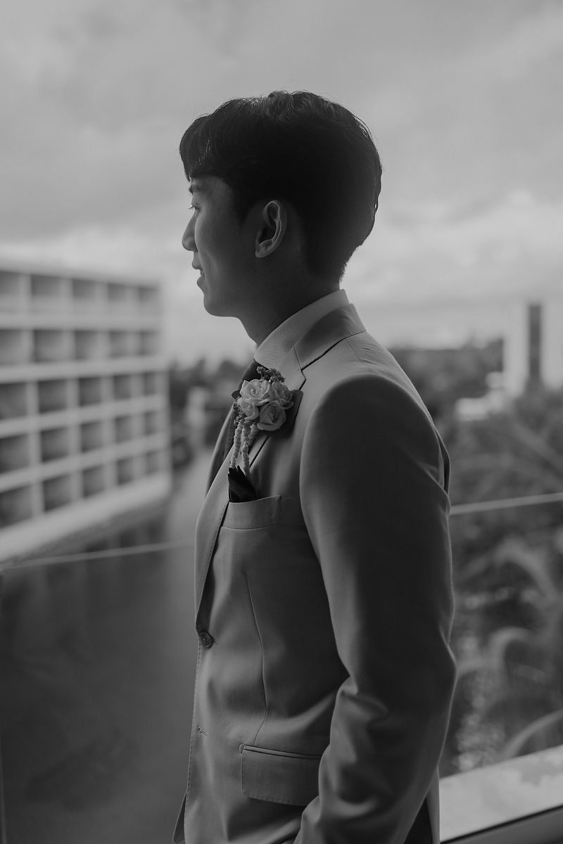 Side profile view of a person in a formal light-colored suit with a boutonniere against a cityscape backdrop in black and white.