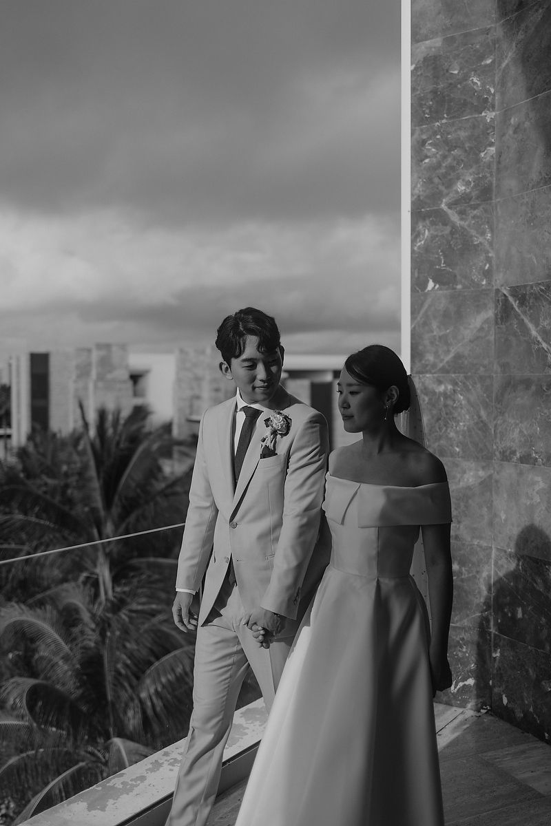 A black and white wedding photograph capturing a couple in formal attire standing on a balcony with palm trees in background.