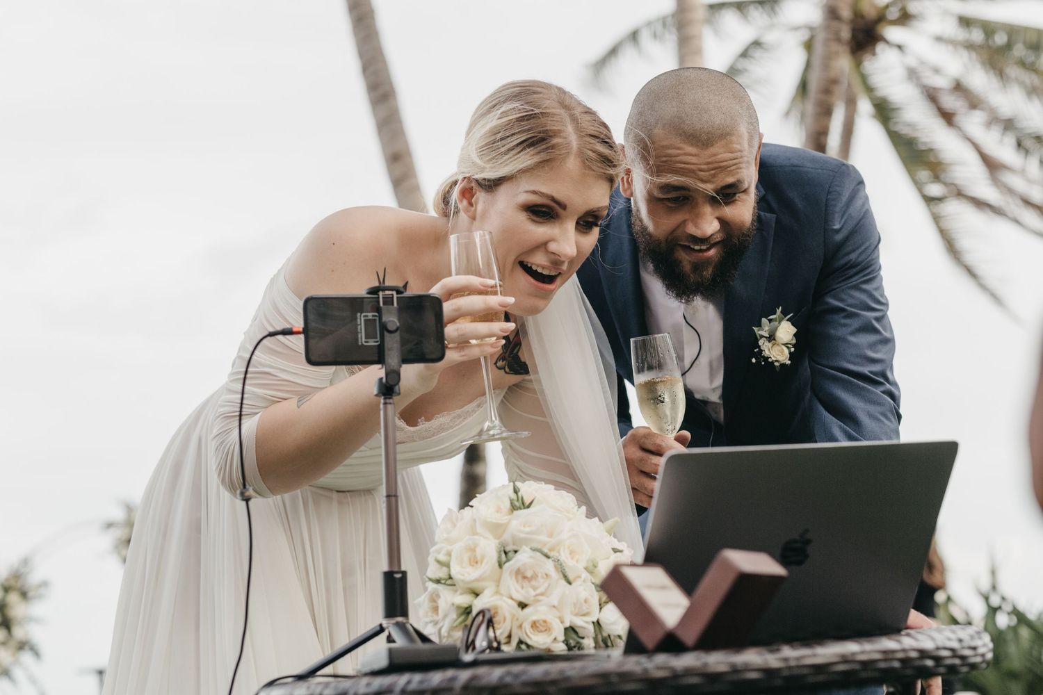 Bride and groom toasting with champagne while video calling on a laptop during an outdoor wedding ceremony.