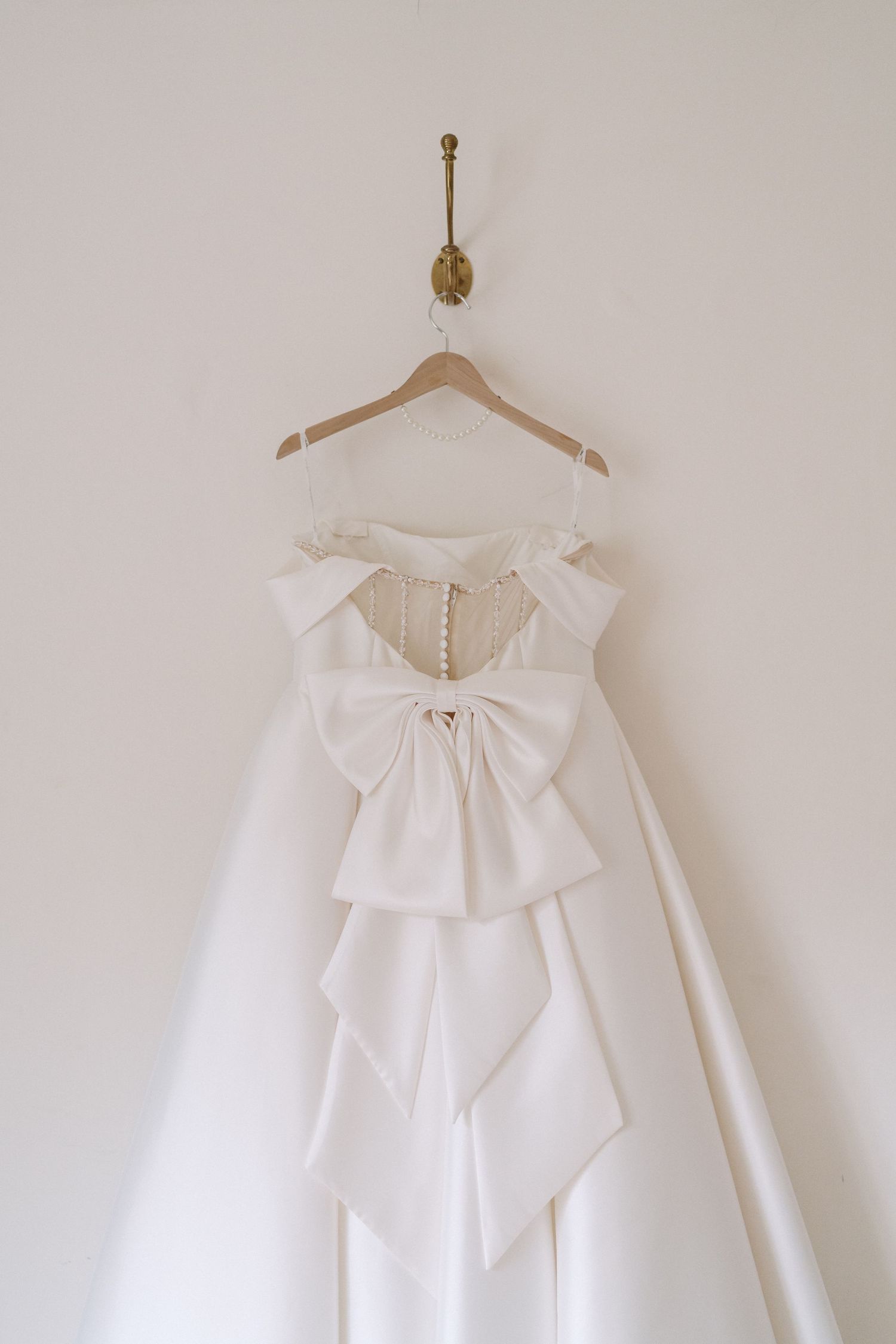 A white wedding dress with a large decorative bow detail hangs elegantly on a wooden hanger against a neutral wall.