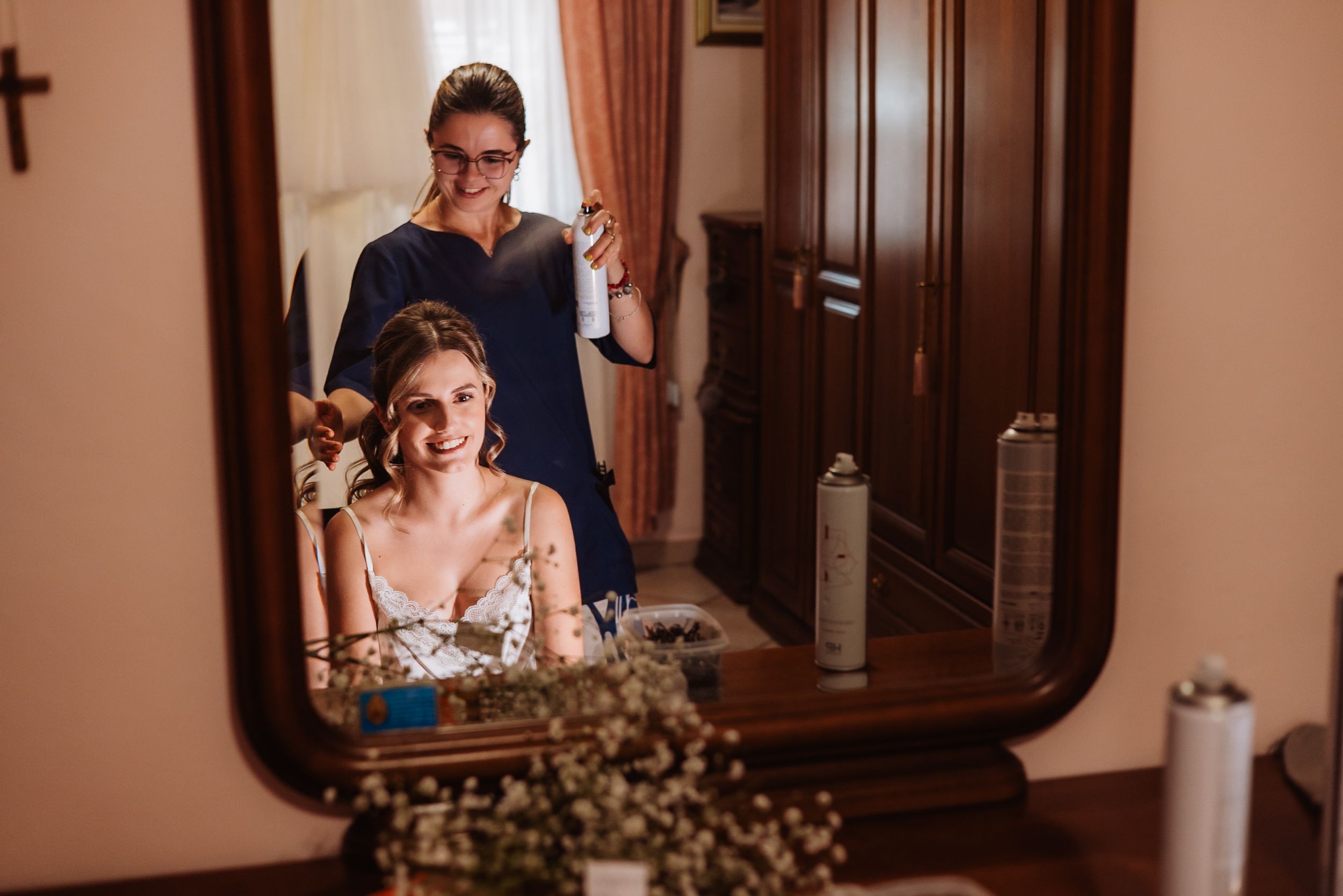 A reflection in an ornate wooden mirror shows people getting ready in a sun-lit room.