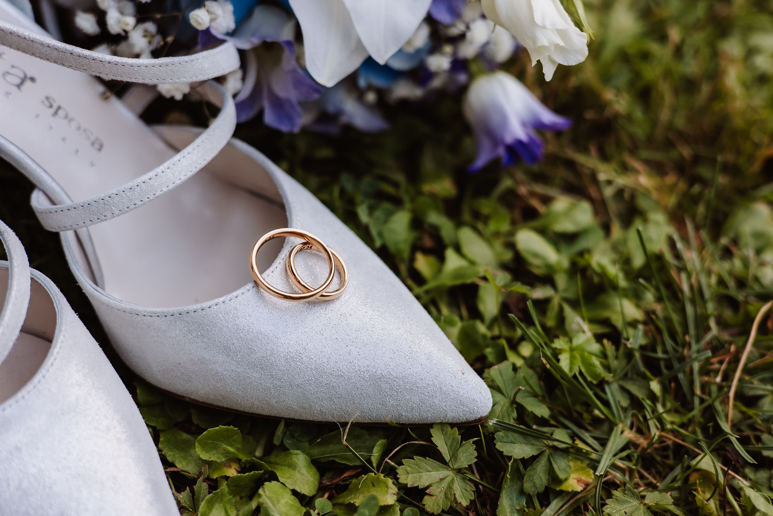 An elegant white bridal shoe sits on grass among delicate purple wildflowers and wedding rings.