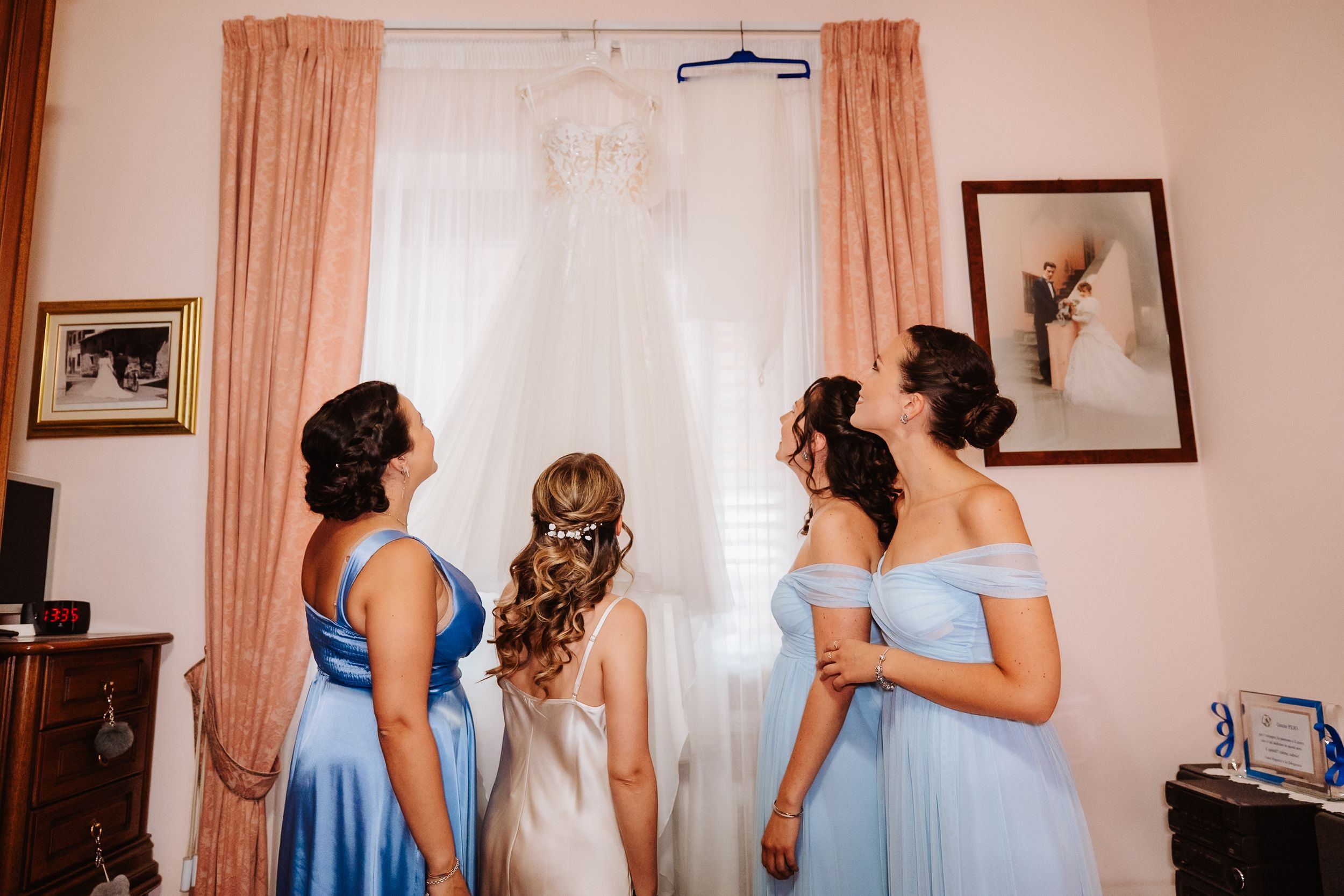 Several women in light blue bridesmaid dresses getting ready in a warmly lit room with peach curtains.