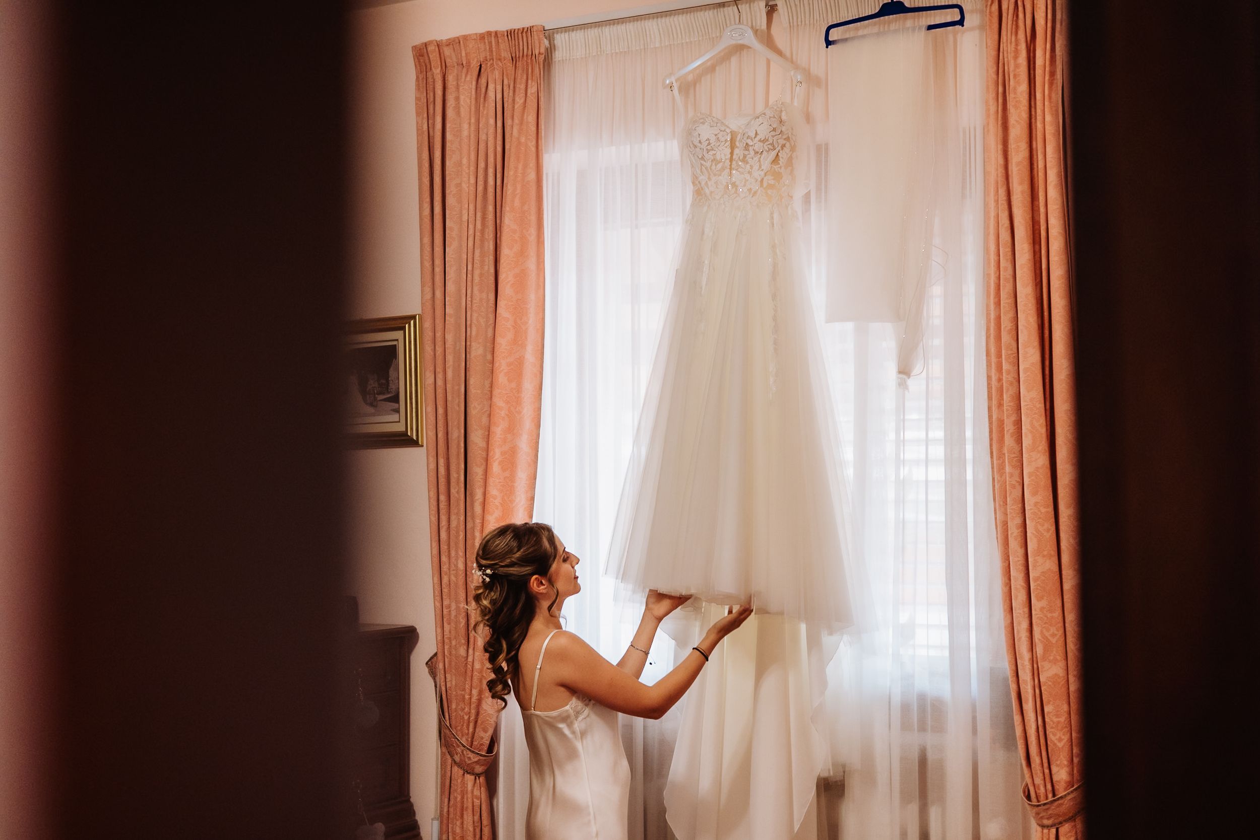 White wedding dress hanging by a window with soft peach curtains in preparation room.