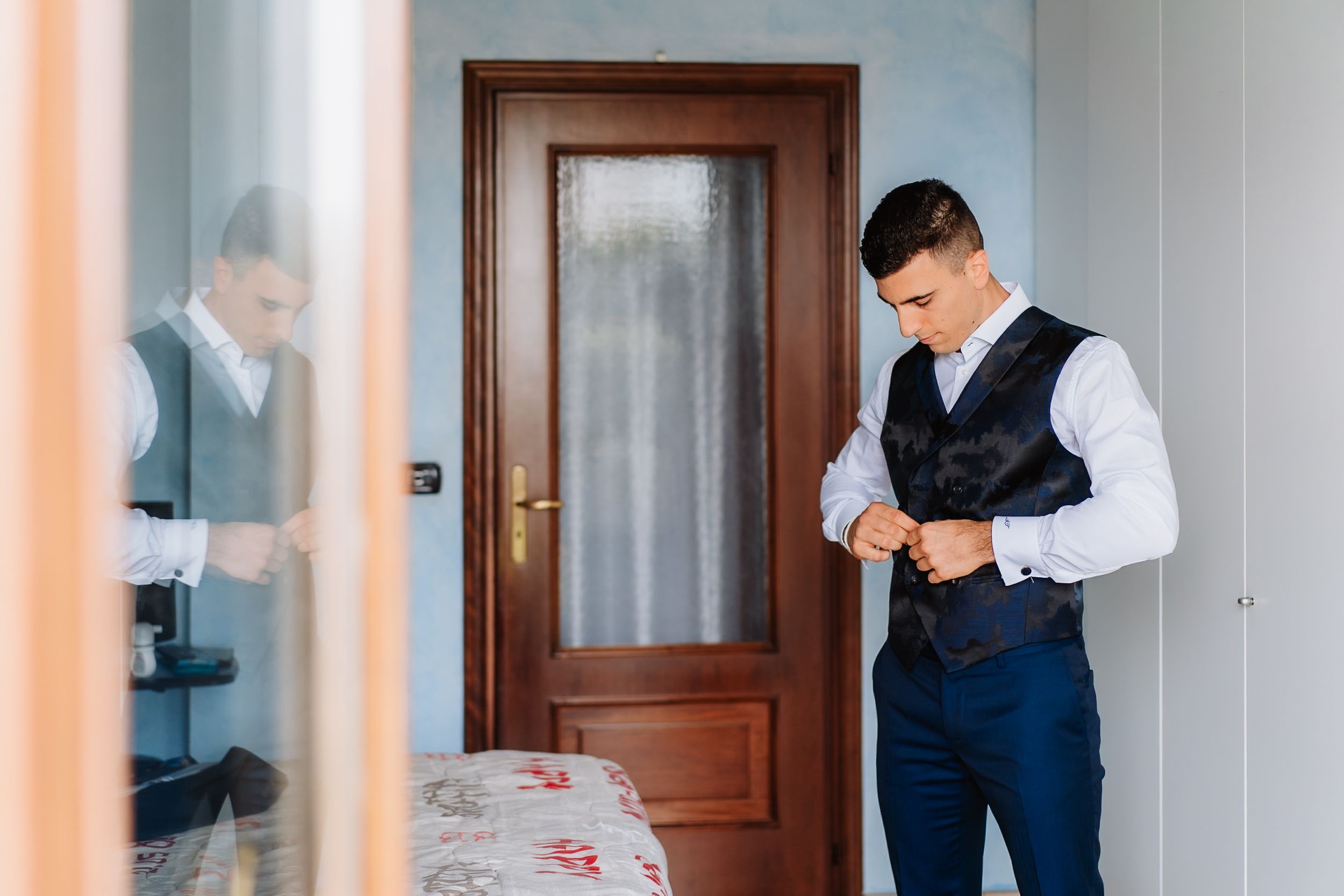 A person in formal attire adjusts a watch while standing in a hallway with wooden doors and orange walls.
