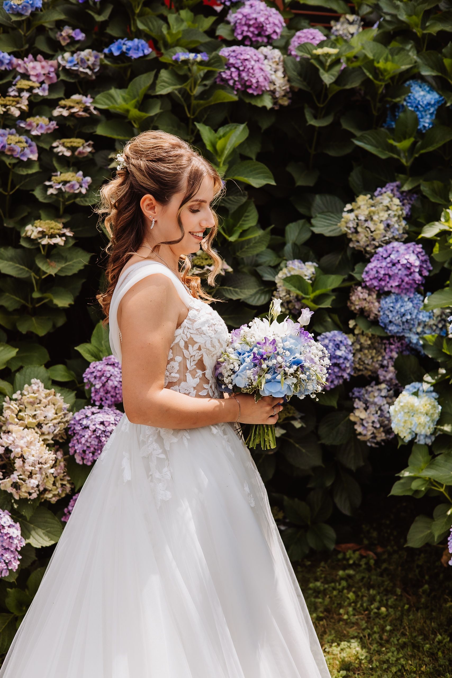 Two white wedding dresses with delicate lace details hang beside vibrant hydrangea flowers in a garden setting.