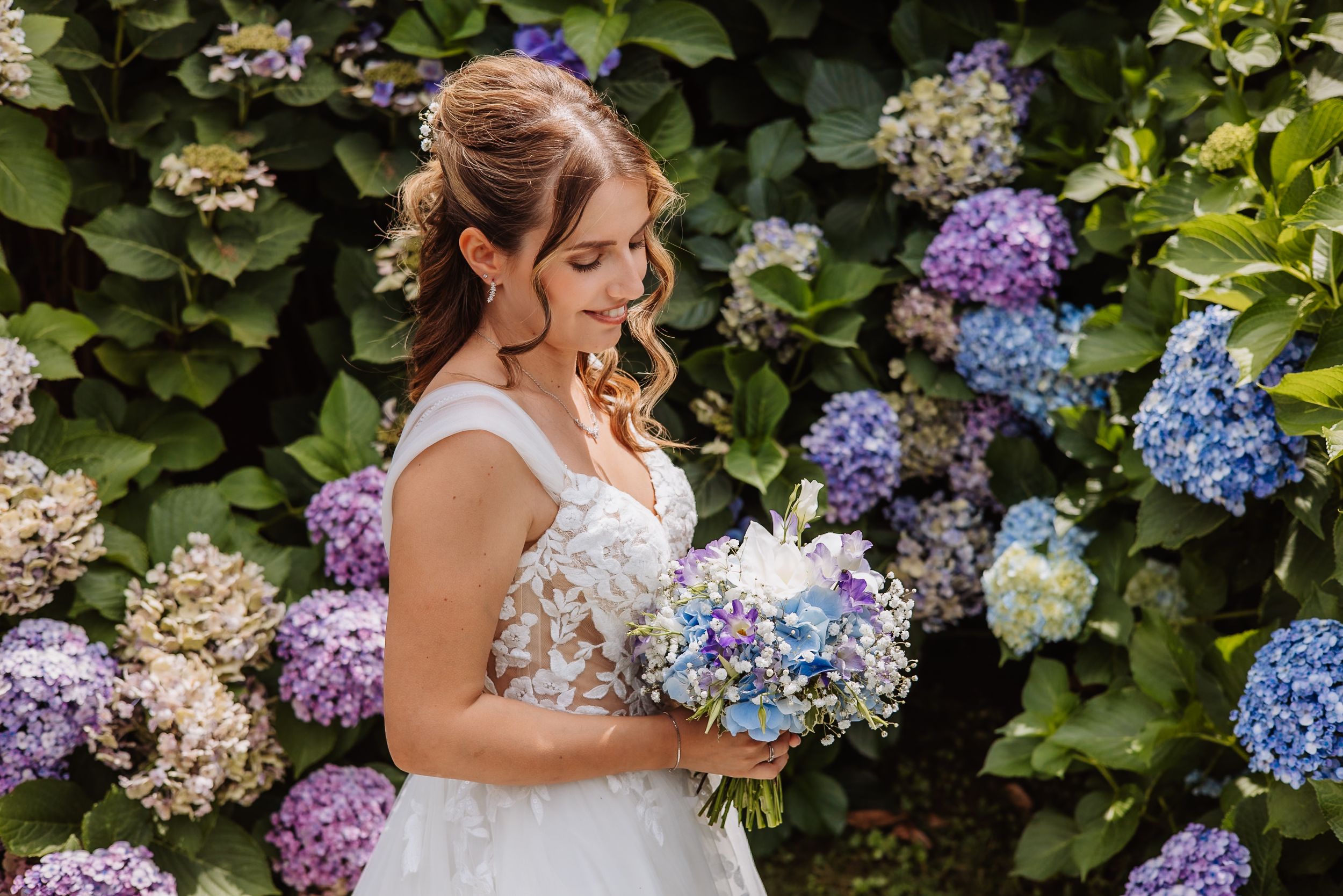 A bride holds a bouquet of purple and blue hydrangeas against a backdrop of lush garden blooms.