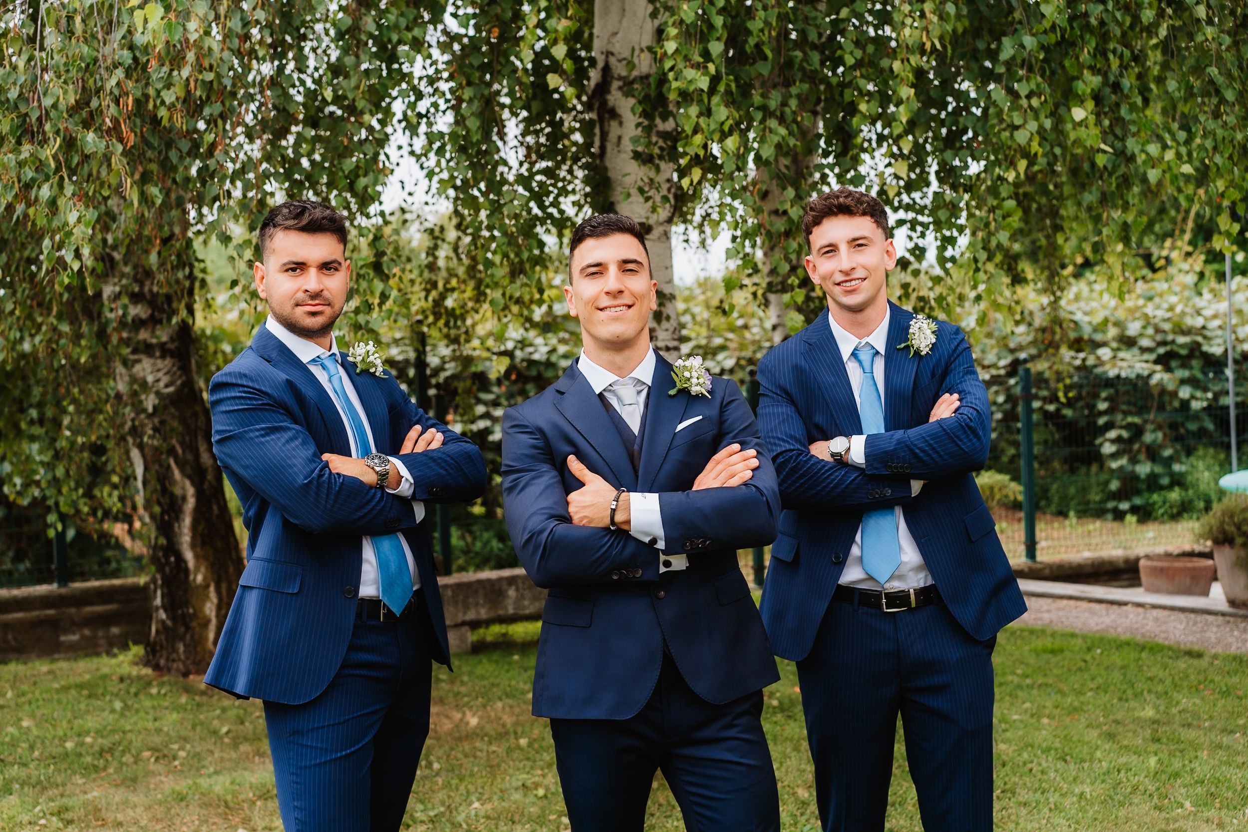 Three groomsmen in matching navy blue suits pose with crossed arms in a garden setting under birch trees.