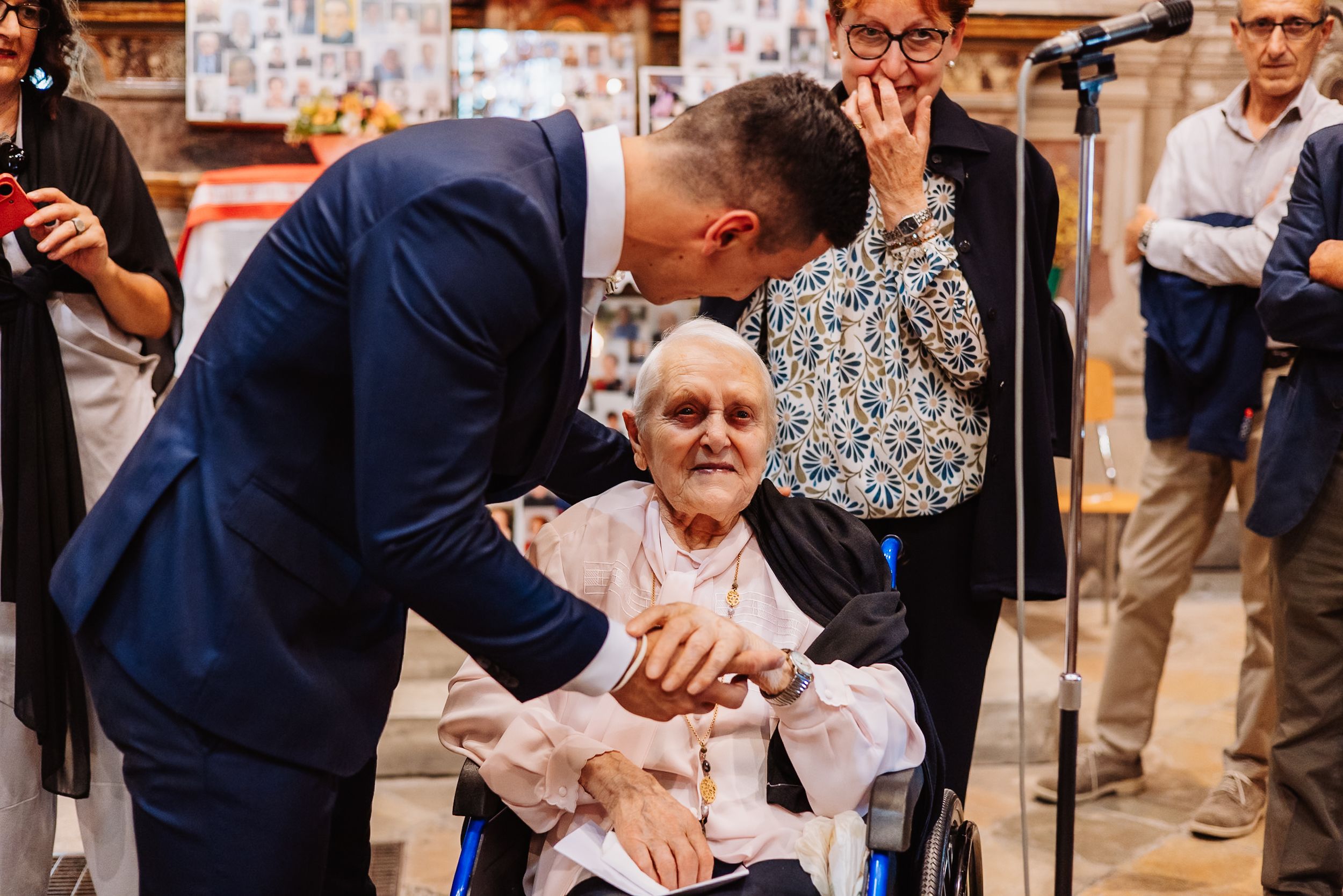A touching moment at a formal gathering shows someone in a navy suit leaning down to greet an elderly person in a wheelchair.