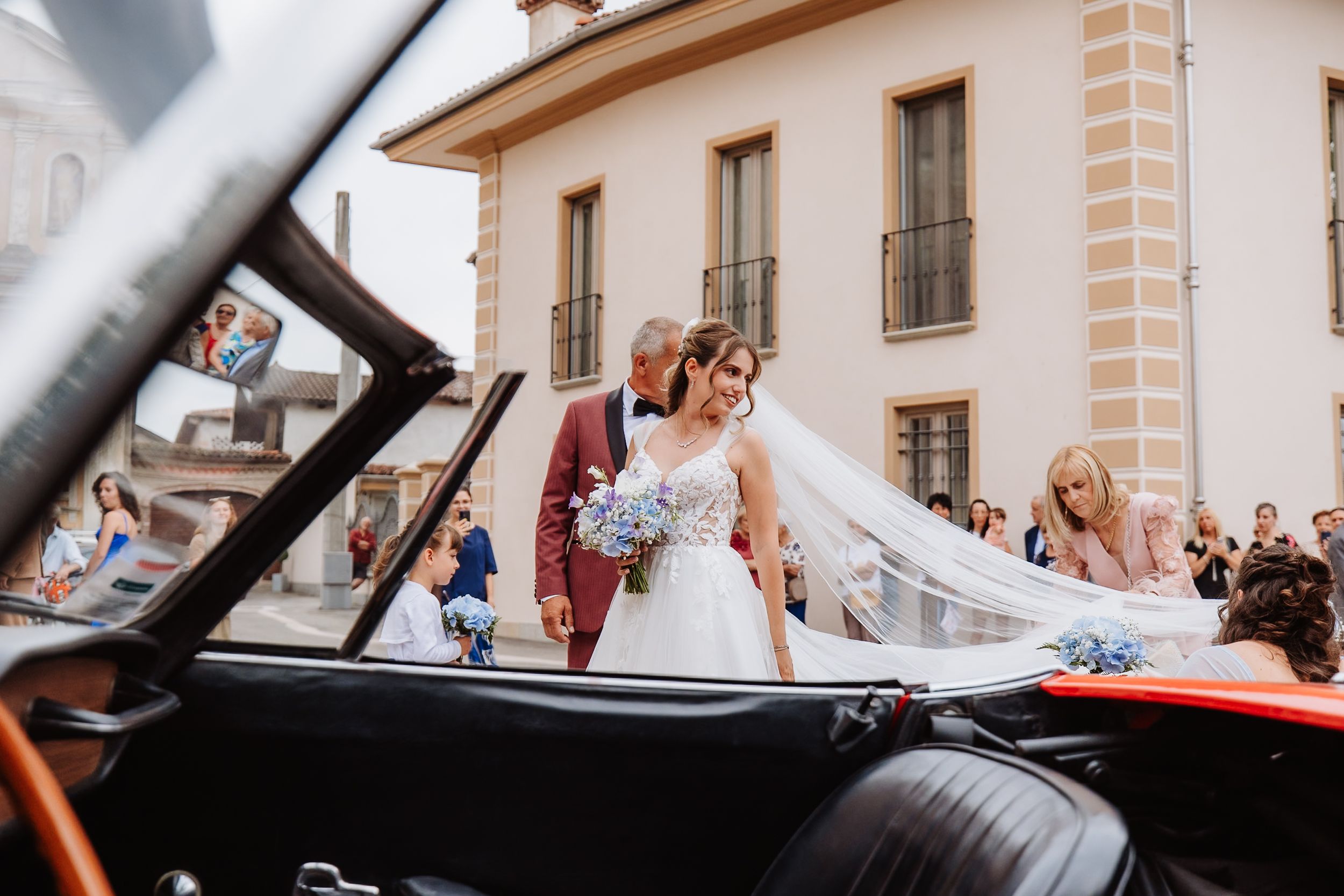 A classic car drives down a street with onlookers gathering along the sides to watch.