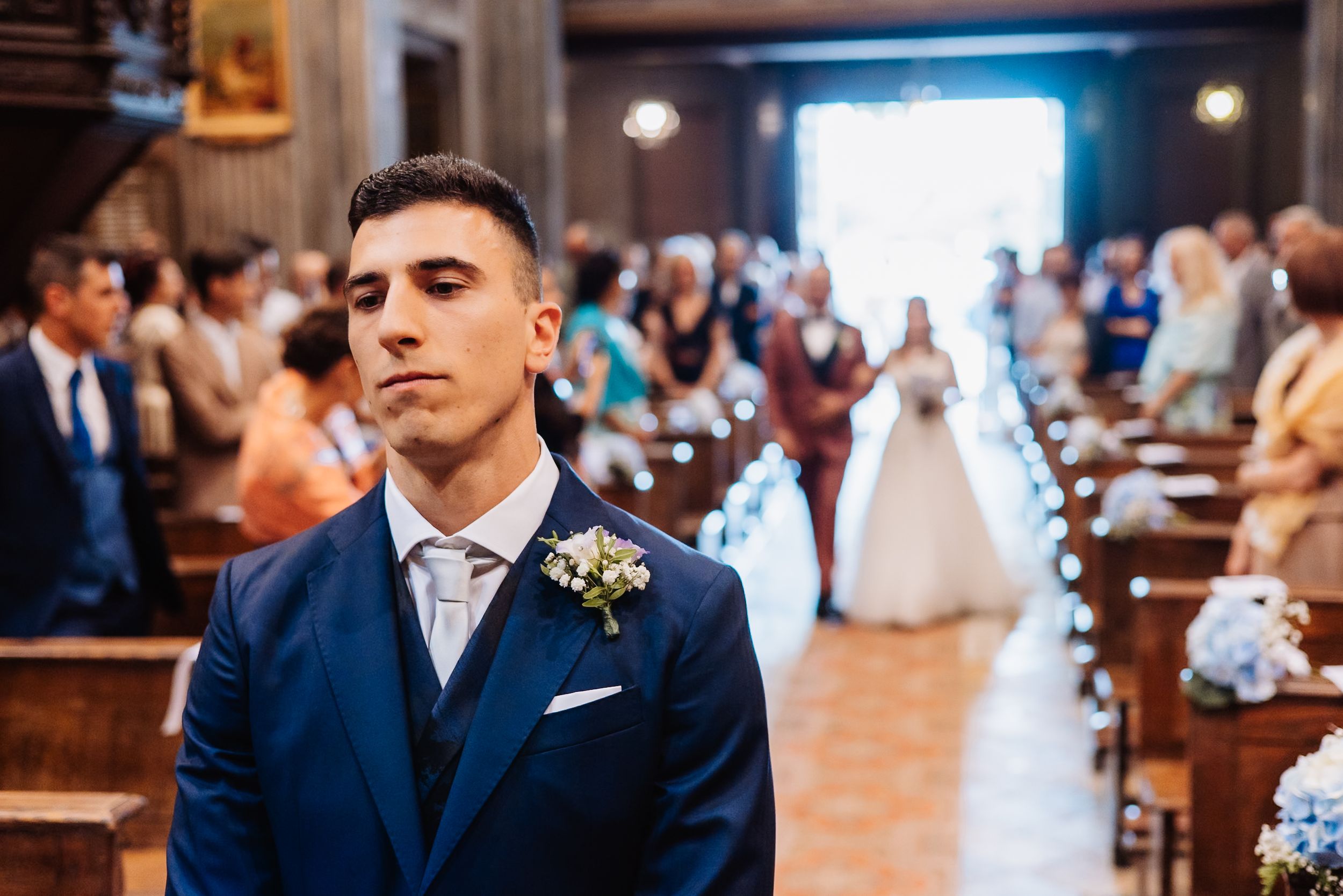 Groom in navy blue suit with boutonniere stands in church pews while bride is visible in background during ceremony.