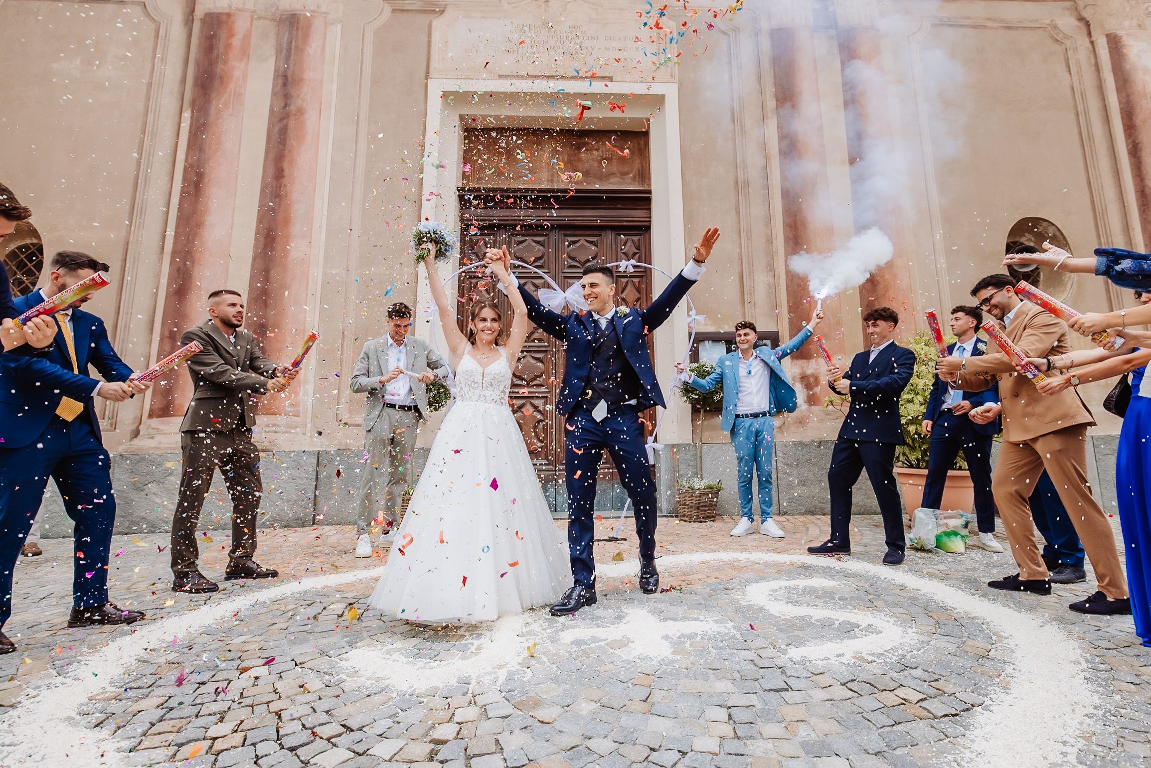 Wedding party celebrating with confetti toss outside a rustic stone building.